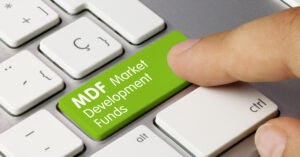 A close-up of a keyboard features a hand pressing a large green key labeled "MDF Market Development Funds," symbolizing the importance of maximizing MDF marketing funds. The surrounding keys are white, highlighting the green key's strategic focus.