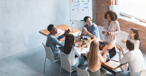 A diverse group of people is seated around a table in a bright conference room, engaged in discussion. A woman is standing, smiling, and holding papers on B2B Marketing Trends 2025, while others sit and clap. A whiteboard with charts is in the background.